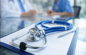 Stetoskop leżący na dokumentacji medycznej na biurku, na pierwszym planie. W tle, delikatnie rozmyci, pracujący personel medyczny w jasnym, nowoczesnym wnętrzu placówki ochrony zdrowia. Naturalne światło dzienne, spokojna atmosfera pracy